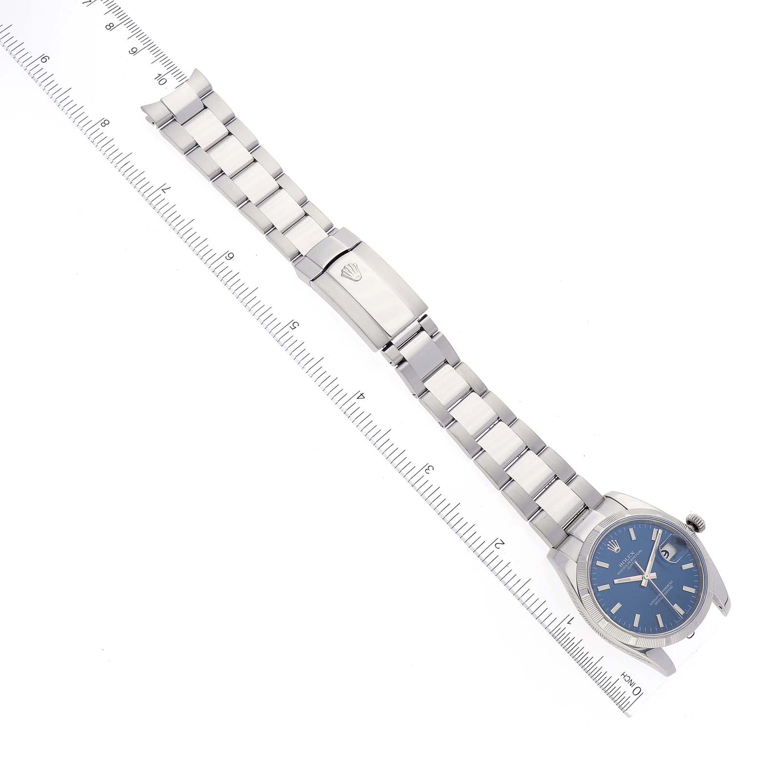 The image shows a Rolex Date 115210 Men's Stainless Steel Blue Dial 115210 Men's Stainless Steel Blue Dial model watch laid flat with a ruler for scale, displaying its bracelet, clasp, and blue dial face.