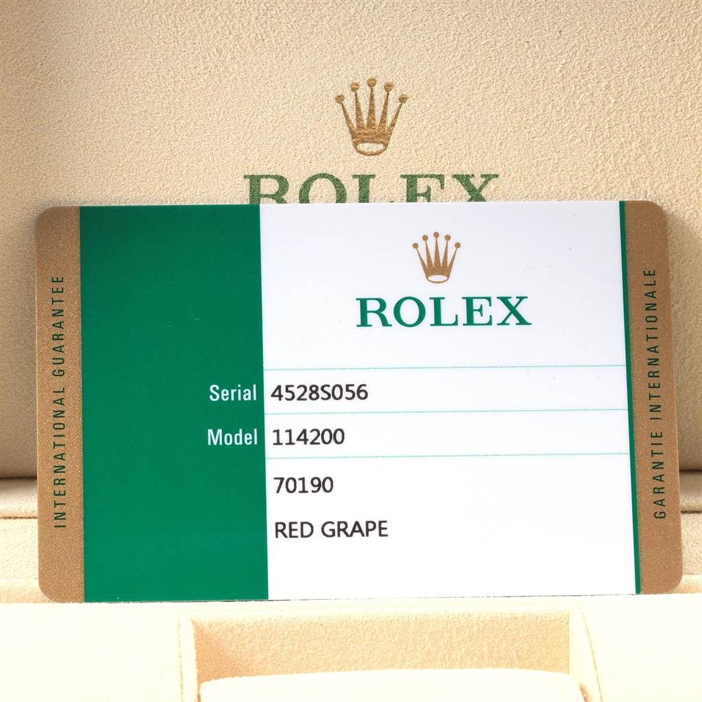 The image shows a warranty card for a Rolex Oyster Perpetual 114200 Men's Stainless Steel Burgundy Dial 114200 Men's Stainless Steel Burgundy Dial model 114200, placed in a beige Rolex box.