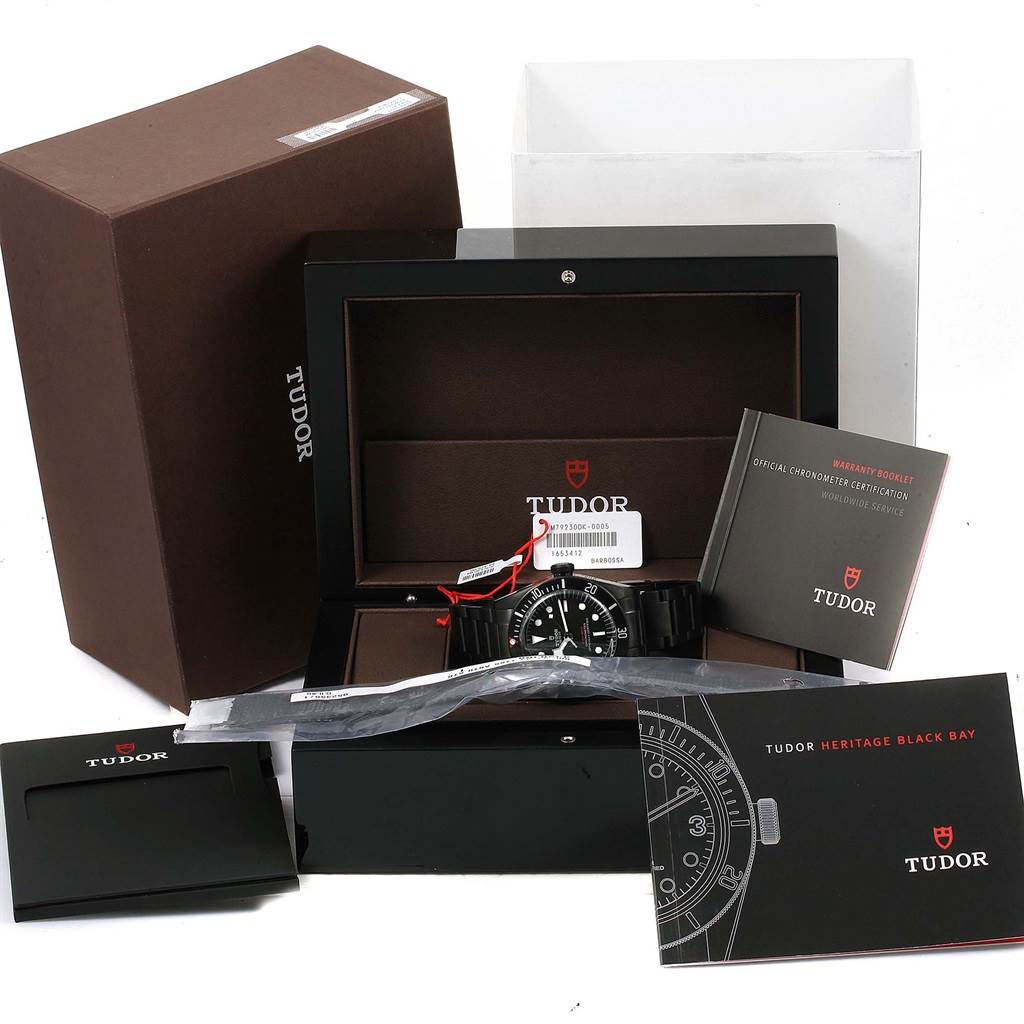 The Tudor Heritage Black Bay 79230DK Men's Stainless Steel Black Dial 79230DK Men's Stainless Steel Black Dial watch is shown with its box, warranty booklet, and accessories viewed from an angle in front.