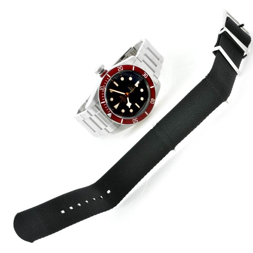 The image shows a Tudor Heritage Black Bay 79220 Men's Stainless Steel Black Dial 79220 Men's Stainless Steel Black Dial watch with a partial view of its metal bracelet and a separate black watch strap.