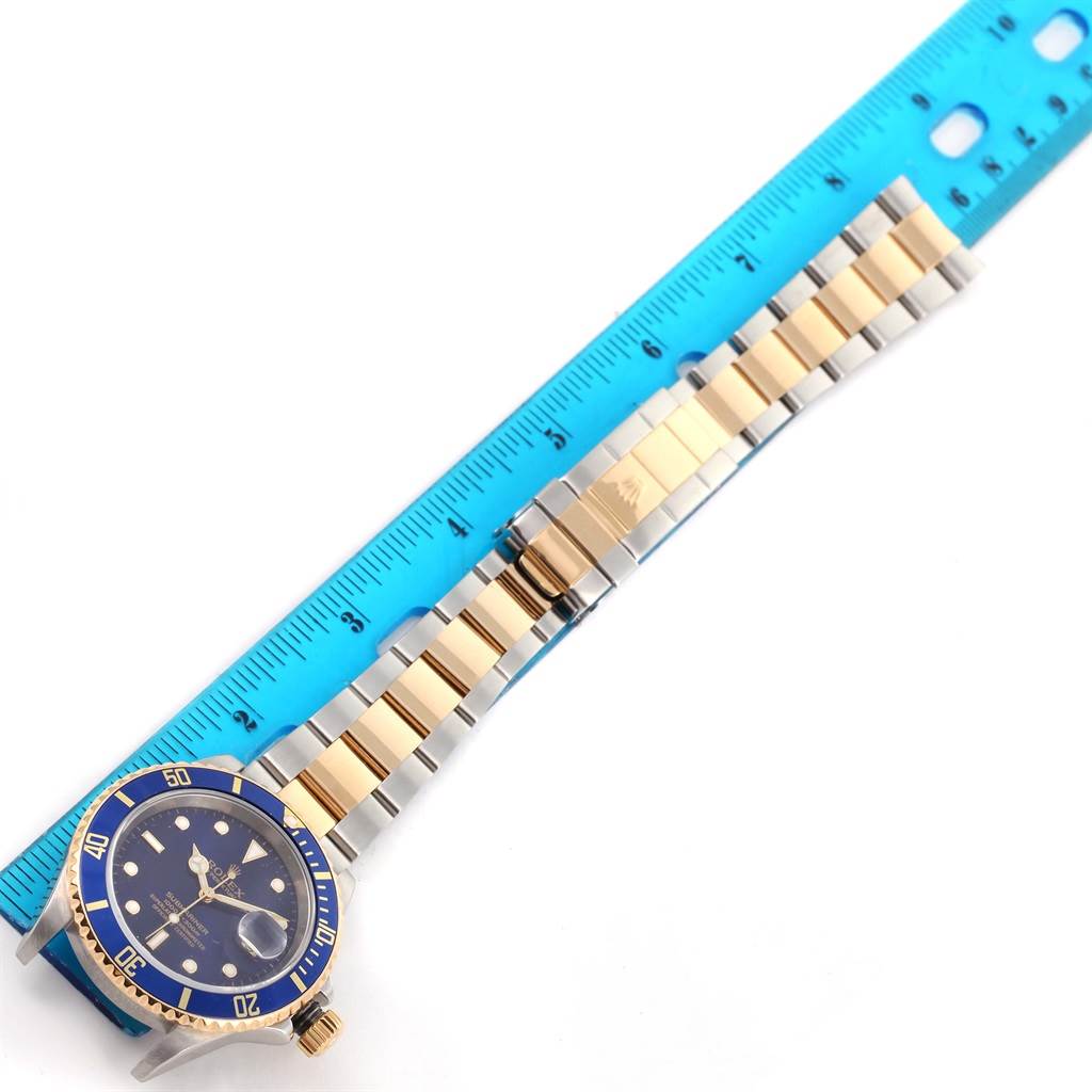 The image shows the Rolex Submariner 16613 Men's Steel and Gold (two tone) Blue Dial 16613 Men's Steel and Gold (two tone) Blue Dial watch lying flat alongside a blue ruler, displaying its blue dial, bezel, and two-tone bracelet.