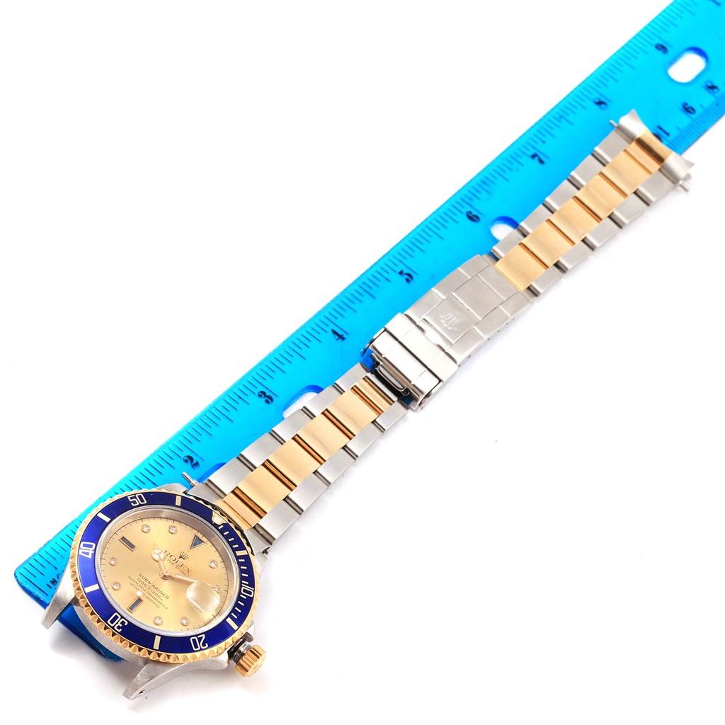 The image shows a Rolex Submariner 16613 Men's Steel and Gold (two tone) Champagne Dial 16613 Men's Steel and Gold (two tone) Champagne Dial watch laying flat with its bracelet against a blue ruler.