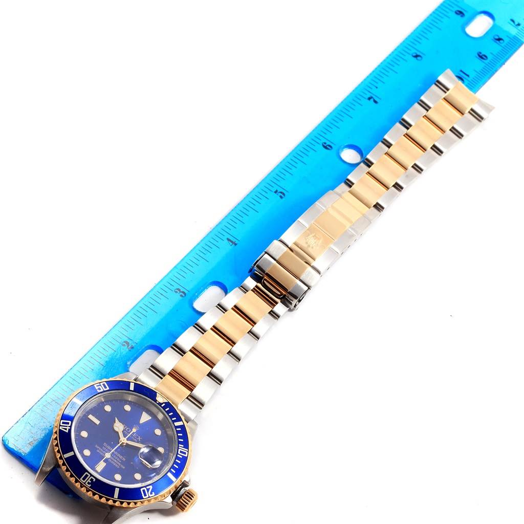 The image shows a Rolex Submariner 16613 Men's Steel and Gold (two tone) Blue Dial 16613 Men's Steel and Gold (two tone) Blue Dial watch placed alongside a ruler, displaying its blue dial, bezel, and two-tone bracelet.