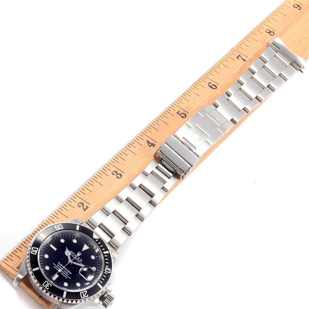 The Rolex Submariner 16610 Men's Stainless Steel Black Dial 16610 Men's Stainless Steel Black Dial watch is shown from a top-down angle with its stainless steel bracelet and a wooden ruler for scale.