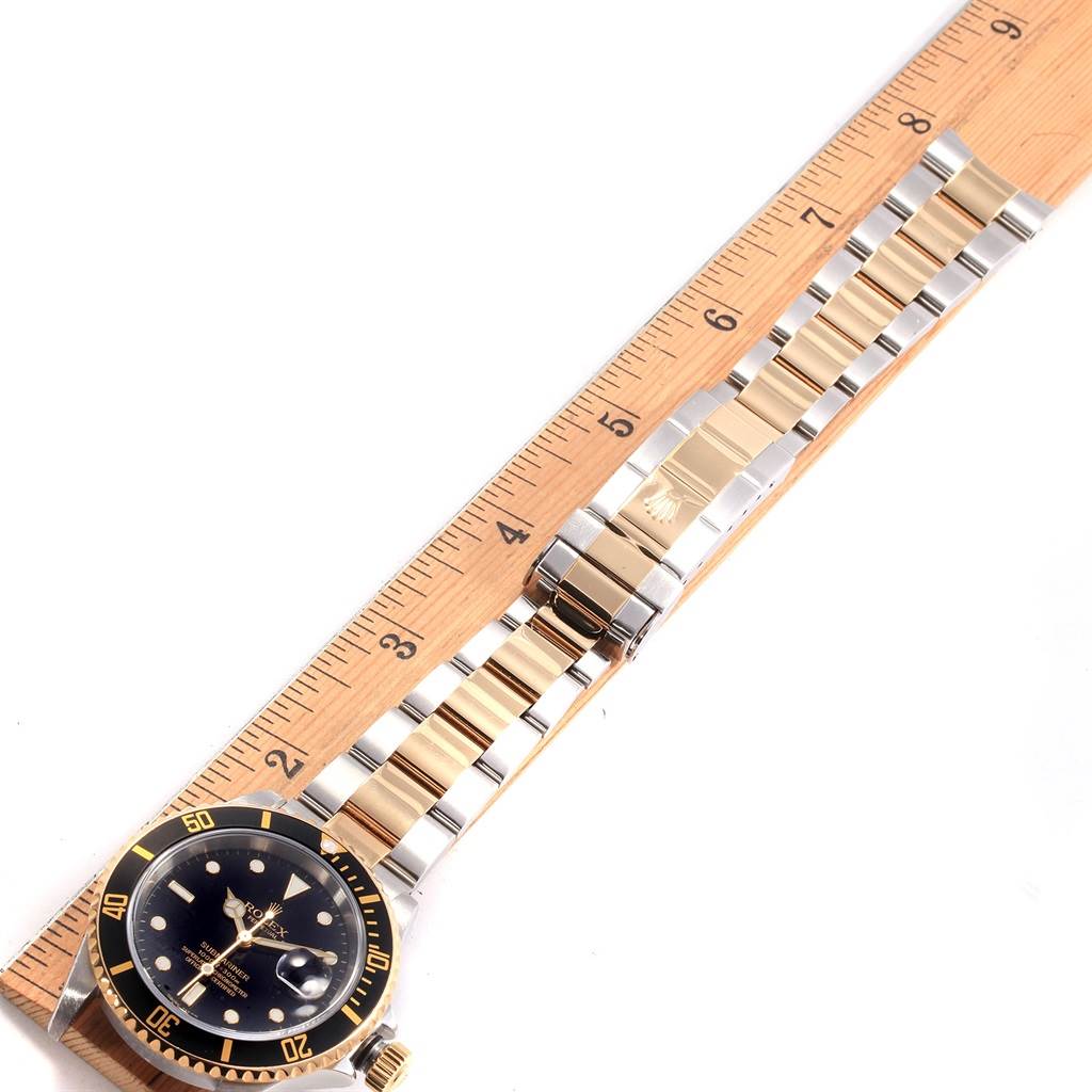 The image shows a Rolex Submariner 16613 Men's Steel and Gold (two tone) Black Dial 16613 Men's Steel and Gold (two tone) Black Dial watch placed on a ruler, highlighting the watch face, bezel, and two-tone metal bracelet.
