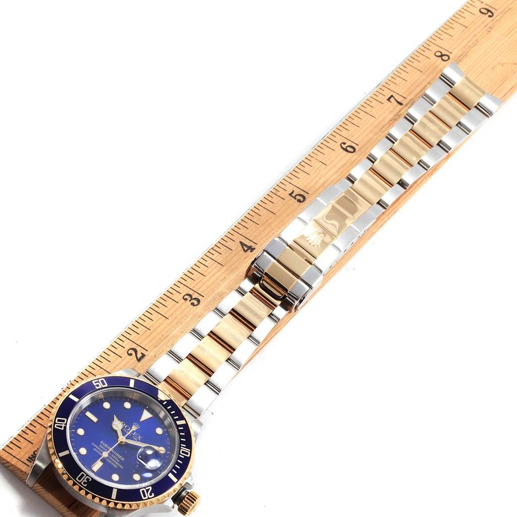 The image shows a Rolex Submariner 16613 Men's Steel and Gold (two tone) Blue Dial 16613 Men's Steel and Gold (two tone) Blue Dial watch laid flat against a wooden ruler, highlighting the face, bezel, and full bracelet length.