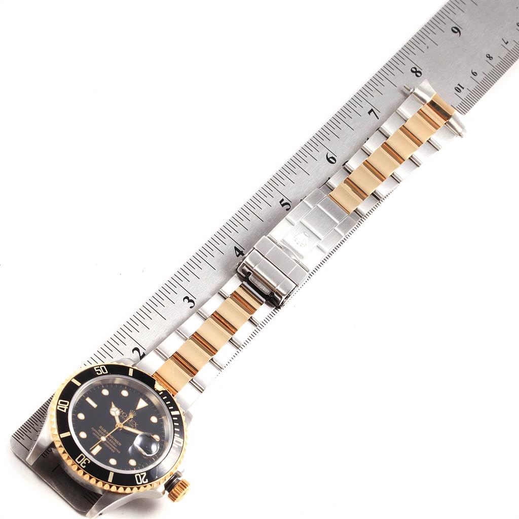 The image shows a Rolex Submariner 16613 Men's Steel and Gold (two tone) Black Dial 16613 Men's Steel and Gold (two tone) Black Dial watch laid flat against a ruler, displaying its face and bracelet.