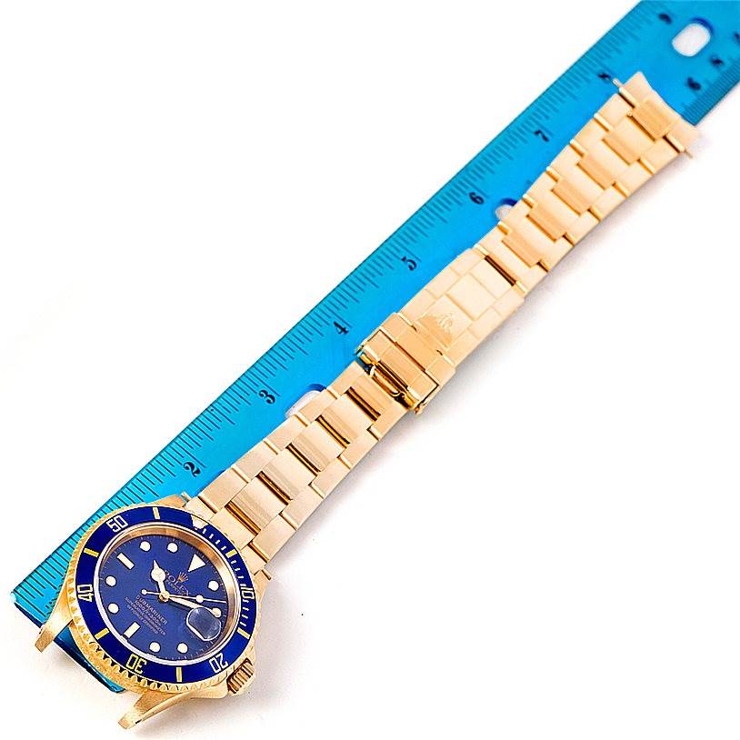 The image shows a top-down view of a Rolex Submariner 16618 Men's Yellow Gold Blue Dial 16618 Men's Yellow Gold Blue Dial watch with a gold bracelet next to a blue ruler.
