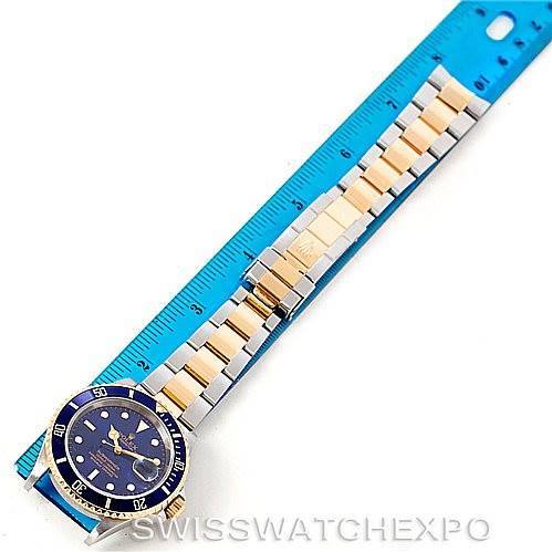 This image shows a Rolex Submariner 16613 Men's Steel and Gold (two tone) Blue Dial 16613 Men's Steel and Gold (two tone) Blue Dial watch laid flat next to a blue ruler, displaying the face and band.