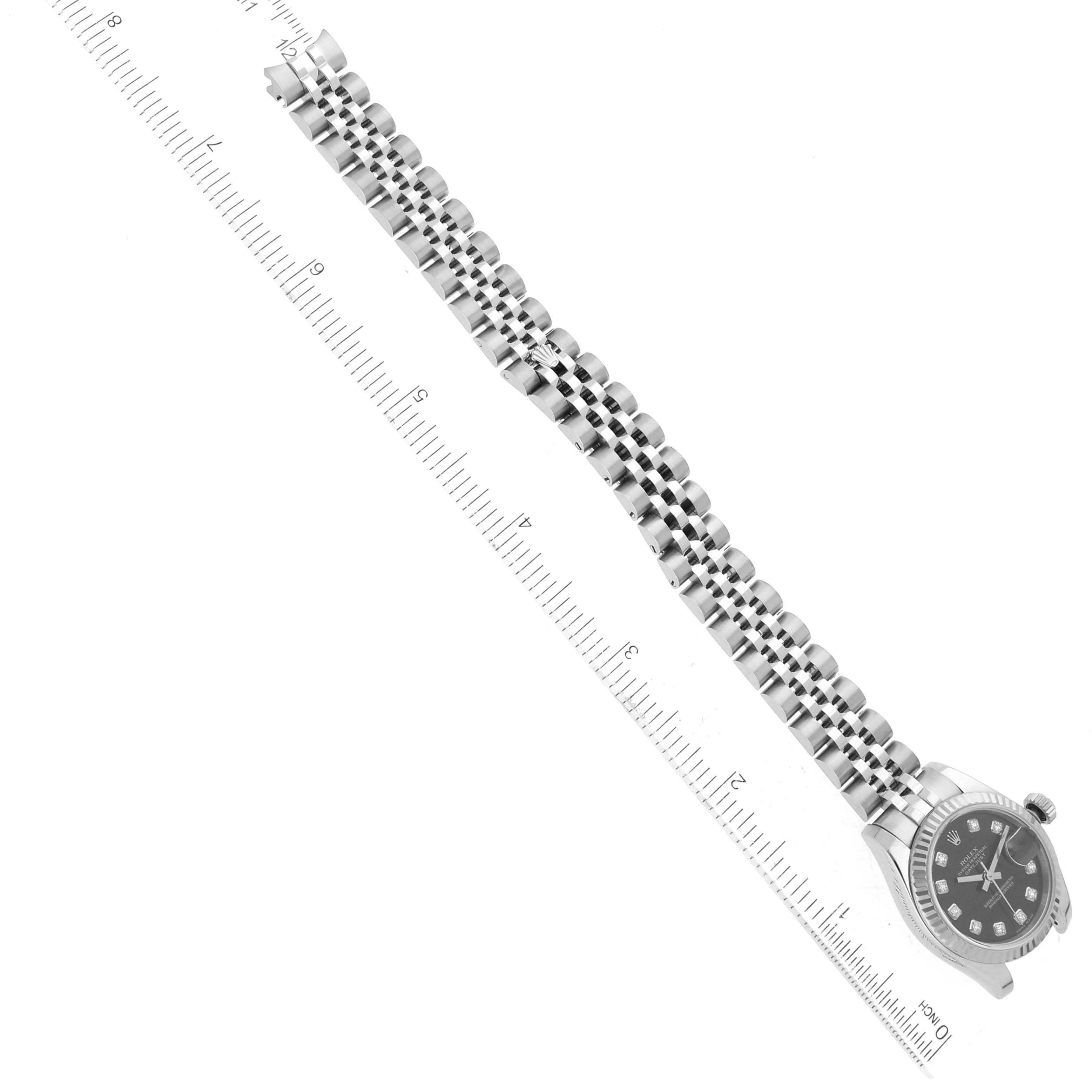 The image shows a diagonal top view of a Rolex Datejust 179174 Women's Stainless Steel Black Dial 179174 Women's Stainless Steel Black Dial watch with a metal bracelet placed against a ruled background.